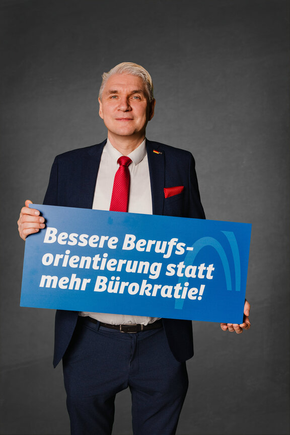 Eine Person steht vor einem neutralen grauen Hintergrund und hält ein großes blaues Schild mit der weißen Aufschrift: "Bessere Berufsoreintierung statt mehr Bürokratie!"