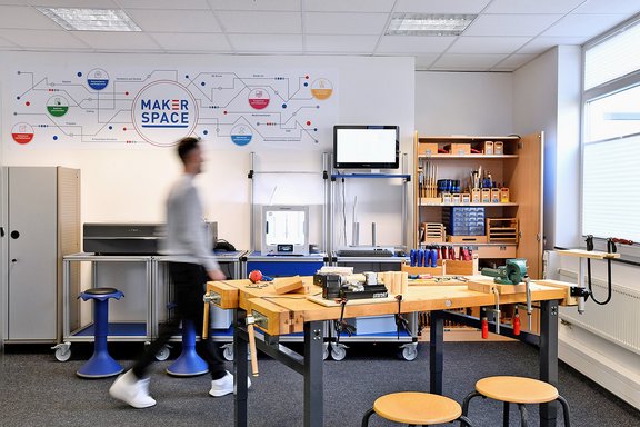 Werkraum mit Werkbank, Werkzeugen, Regalen und Monitor, an der Wand Schriftzug 'MAKER SPACE' mit vernetzten Symbolen, Person in Bewegung unscharf.