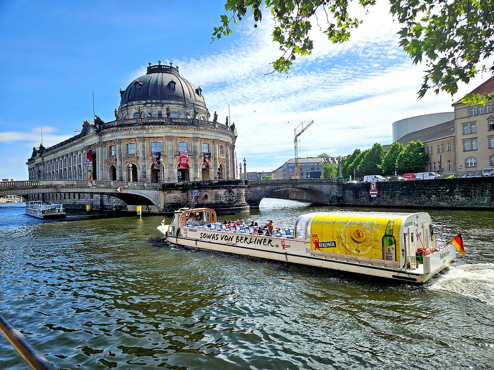 Ein Ausflugsboot mit Passagieren fährt auf einem Fluss vorbei an der Museumsinsel in Berlin. Im Hintergrund ist das Bode-Museum sichtbar.