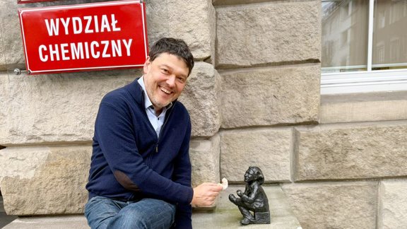 Mann in blauer Jacke und Jeans sitzt auf einer Steinbank neben einer kleinen bronzenen Statue eines sitzenden Kindes, rote Schilder mit weißer Aufschrift 'POLITECHNIKA WROCŁAWSKA' und 'WYDZIAŁ CHEMICZNY' an der Wand.