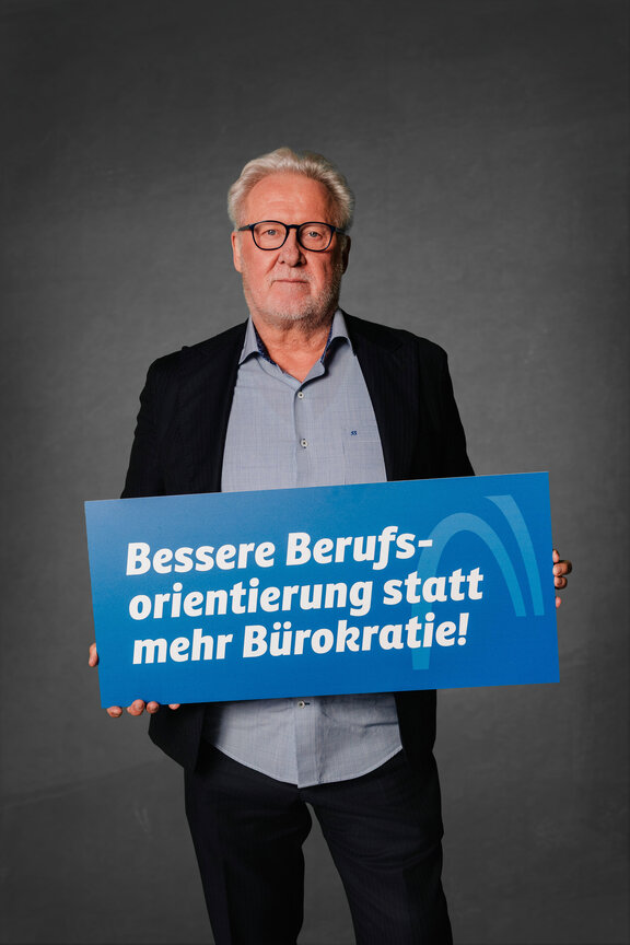 Eine Person steht vor einem neutralen grauen Hintergrund und hält ein großes blaues Schild mit der weißen Aufschrift: "Bessere Berufsorientierung statt mehr Bürokratie!"
