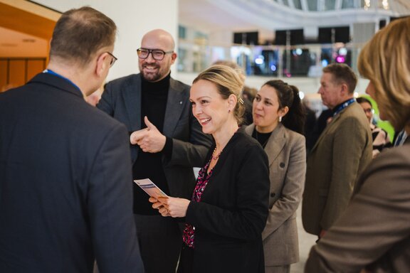 Zu sehen ist eine Gruppe von Menschen, die sich unterhalten. In der Bildmitte befindet sich eine mittelalte blonde Frau in einem schwarzen Sakko. Neben ihre ein Mann mit Glatze und Brille, der einen schwarzen Rollkragen und ebenfalls ein schwarzes Sakko trägt. Es handelt sich um die IHK-Hauptgeschäftsführerin Manja Schreiner und dem Generalsekretär Michael Biel.