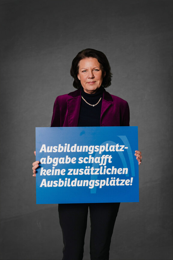 Eine Person steht vor einem neutralen grauen Hintergrund und hält ein großes blaues Schild mit der weißen Aufschrift: "Die Strafabgabe schwächt den Wirtschaftsstandort!"