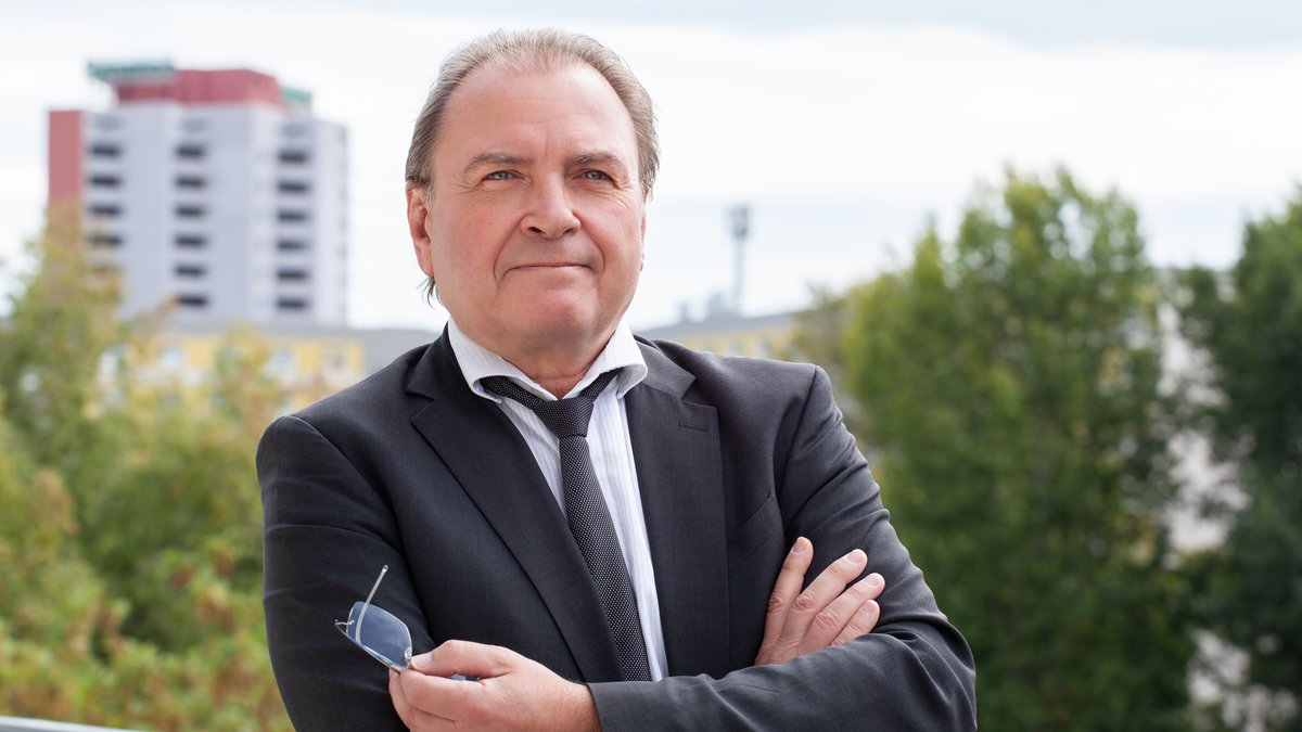 Mann in dunklem Anzug mit weißem Hemd und Krawatte steht mit verschränkten Armen auf einem Balkon vor Bäumen und Hochhäusern.