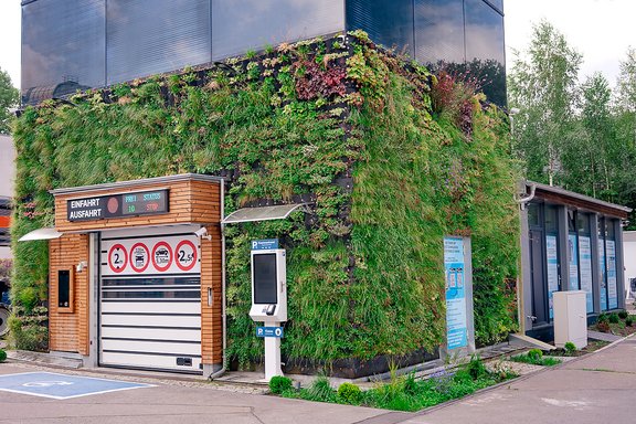 Eine moderne Parkhausfassade mit vertikaler Begrünung. Links ein Zugang mit drei roten Verkehrssymbolen: '2', '2.5', '3'. Rechts ein Ticketautomat.