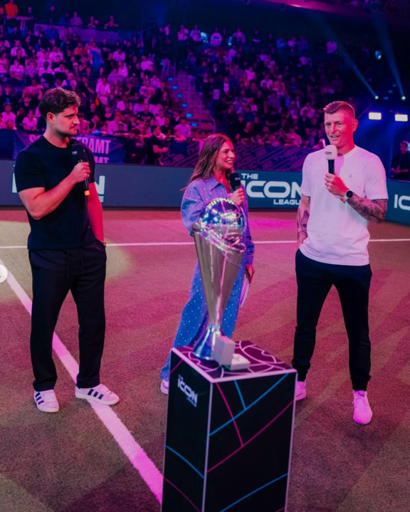 Drei Personen mit Mikrofonen stehen auf einem Spielfeld neben einem großen Pokal, umgeben von Publikum in einer Arena mit bunter Beleuchtung.