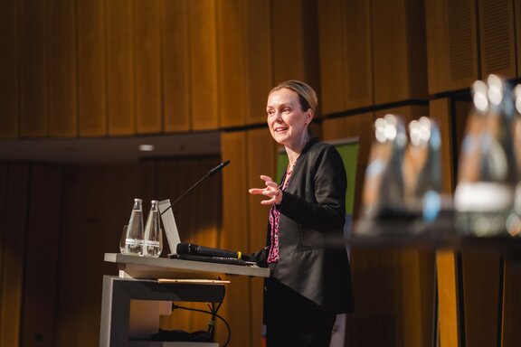 Zu sehen ist eine mittelalte blonde Frau in einem dunklen Sakko. Sie steht auf einer Bühne und spricht in ein Mikrofon. Es handelt sich um die Hauptgeschäftsführerin der IHK Berlin Manja Schreiner.