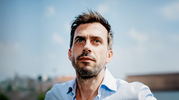 Mann mit offenem hellblauem Hemd vor unscharfem Himmel und Gebäuden im Hintergrund.