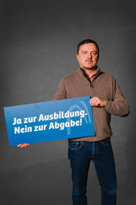 Eine Person steht vor einem neutralen grauen Hintergrund und hält ein großes blaues Schild mit der weißen Aufschrift: "Ja zur Ausbildung. Nein zur Abgabe!"