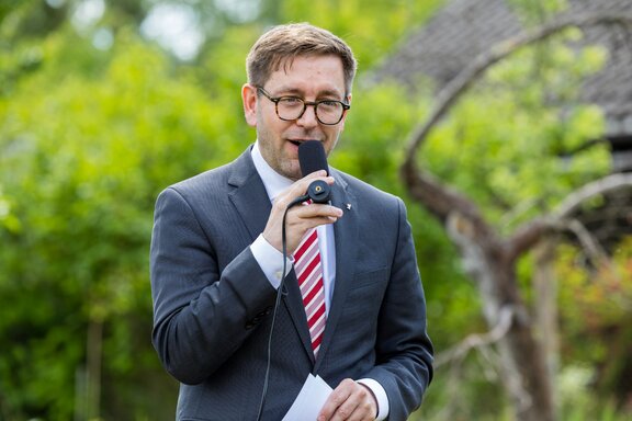 Zu sehen ist ein mittelalter Mann mit Brille und Anzug, der in eine Mikrofon spricht. Es handelt sich um Staatssekretär Andreas Kraus.