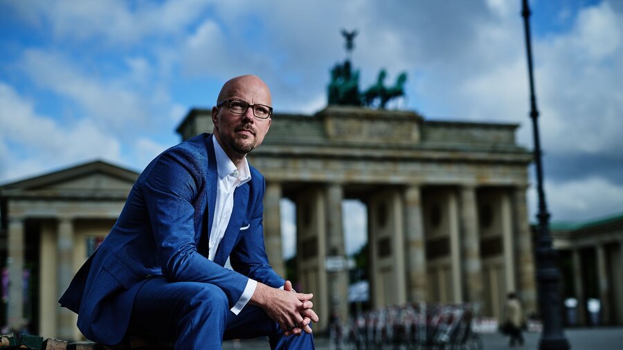 Zu sehen ist ein Mann in blauem Anzug mit Brille und Glatze. Er sitzt vorgebeugt auf einer Bank, im Hintergrund das Brandenburger Tor. Es handelt sich um Sebastian Stietzel.
