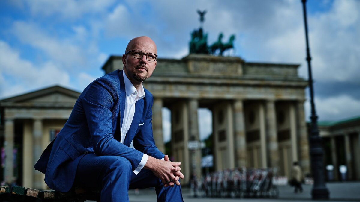 Zu sehen ist ein Mann in blauem Anzug mit Brille und Glatze. Er sitzt vorgebeugt auf einer Bank, im Hintergrund das Brandenburger Tor. Es handelt sich um Sebastian Stietzel.
