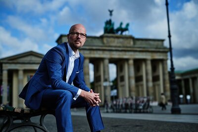 Zu sehen ist ein Mann in blauem Anzug mit Brille und Glatze. Er sitzt vorgebeugt auf einer Bank, im Hintergrund das Brandenburger Tor. Es handelt sich um Sebastian Stietzel.