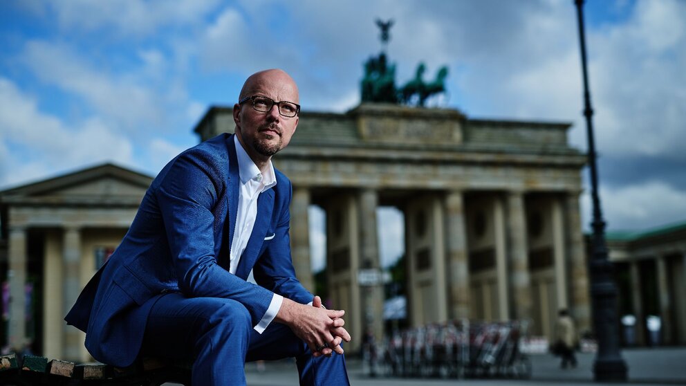 Zu sehen ist ein Mann in blauem Anzug mit Brille und Glatze. Er sitzt vorgebeugt auf einer Bank, im Hintergrund das Brandenburger Tor. Es handelt sich um Sebastian Stietzel.