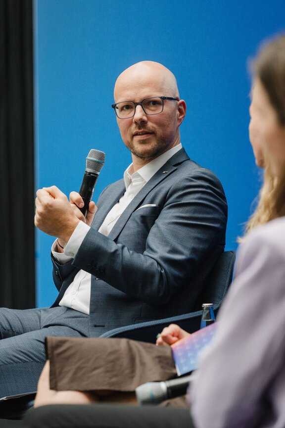 Zu sehen ist ein Mann mit Glatze und Brille im Anzug. Er spricht in ein Mikrofon und gestikuliert dabei. Es handelt sich um Sebastian Stietzel.