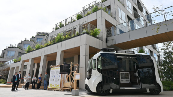 Ein autonomer Kleinbus steht vor einem modernen Wohnhaus. Daneben stehen sechs Menschen in einer Gruppe. 
