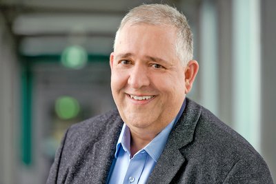 Älterer Mann mit grauem Haar, trägt hellblaues Hemd und grauen Blazer, steht in einem Flur mit unscharfem Hintergrund.