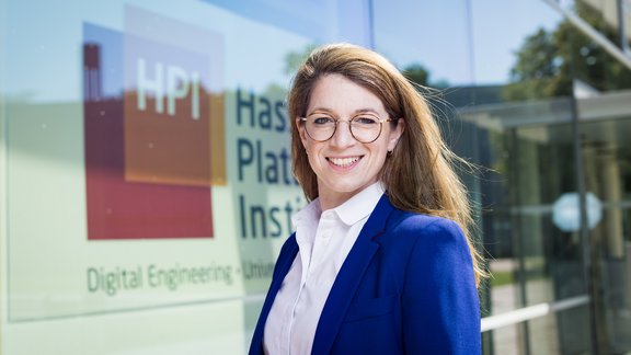 Frau mit langen blonden Haaren trägt weißen Hemdkragen und blauen Blazer, steht vor Glasfassade mit HPI-Logo und Schriftzug.