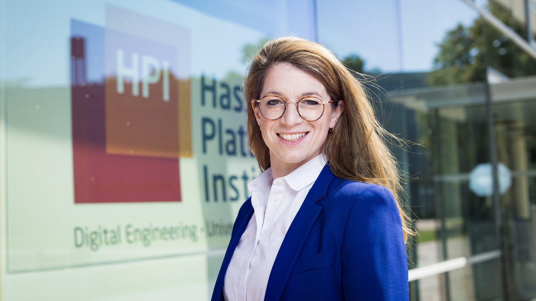 Frau mit langen blonden Haaren trägt weißen Hemdkragen und blauen Blazer, steht vor Glasfassade mit HPI-Logo und Schriftzug.