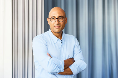 Mann mit hellblauem Hemd steht mit verschränkten Armen vor grauen Vorhängen.