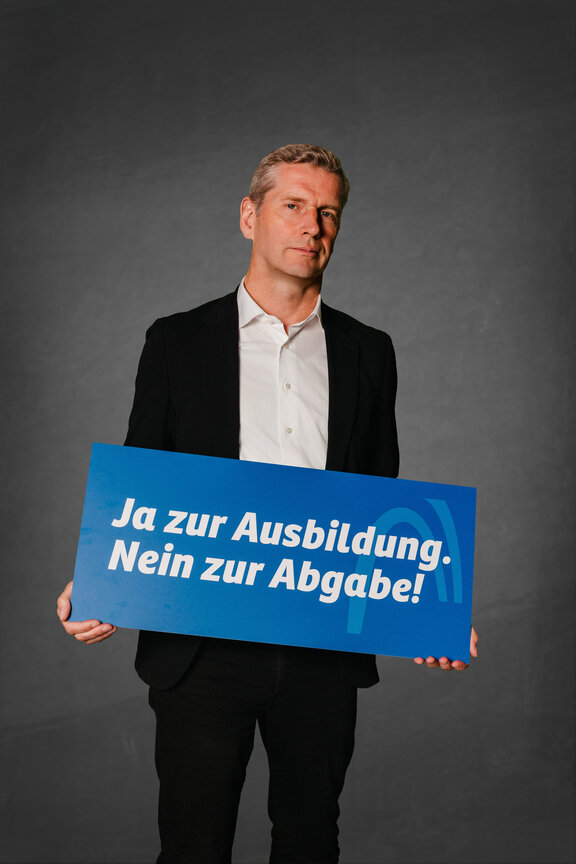 Eine Person steht vor einem neutralen grauen Hintergrund und hält ein großes blaues Schild mit der weißen Aufschrift: "Ja zur Ausbildung. Nein zur Abgabe!"