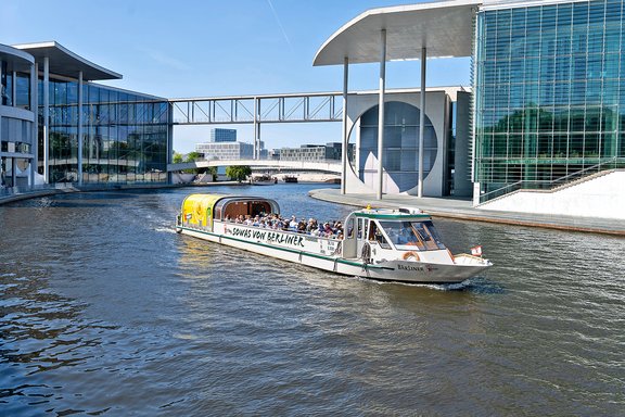 Ein Ausflugsboot mit gelbem Dach fährt auf einem Wasserweg zwischen modernen Gebäuden. Auf dem Boot sind Passagiere zu sehen.
