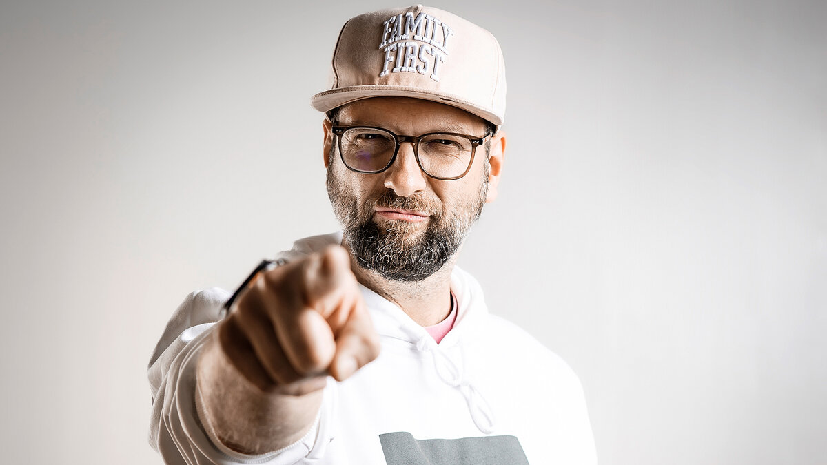 Person mit Baseballkappe und weißem Kapuzenpullover zeigt mit ausgestrecktem Arm direkt auf die Kamera.