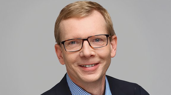 Mann mit kurzen blonden Haaren trägt dunklen Anzug und blau-weiß kariertes Hemd vor grauem Hintergrund.