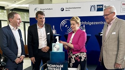 Drei Männer und eine Frau stehen u eine dreistöckige blaue Torte, welche von der Frau, die ein pinkes Sakko trägt, angeschnitten wird.