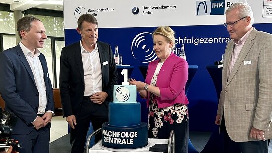 Drei Männer und eine Frau stehen u eine dreistöckige blaue Torte, welche von der Frau, die ein pinkes Sakko trägt, angeschnitten wird.