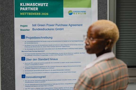Zu sehen ist eine Frau mit kurzen blonden Haaren, die vor einer Infotafel steht und zur Seite blickt.