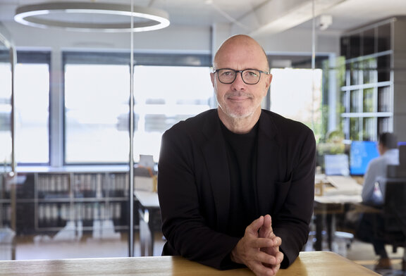 Ein Mann mit Brille steht an einem Stehschreibtisch in einem Büro und lächelt in die Kamera. Es ist Thomas Bestgen.  