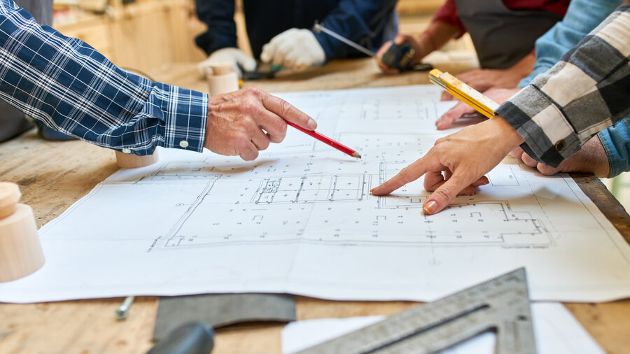 Auf einem Holztisch liegt ein Bauplan. Eine Hand zeigt auf den Bauplan und die andere Hand zeigt mit einem Stift auf eine andere Stelle.