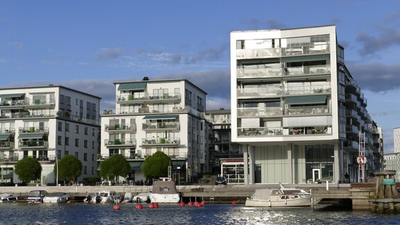 Drei moderne Mehrfamilienhäuser an einer Uferpromenade. Auf dem Wasser schwimmen kleine Boote. 