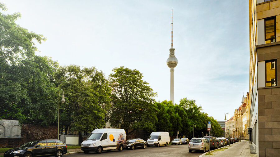 Zu sehen ist eine befahrene Straße. Am linken Straßenrand stehen viele Bäume, auf der Anderen Seite stehen Wohnhäuser. Im Hintergrund ist der Fernsehturm in Berlin zu sehen.
