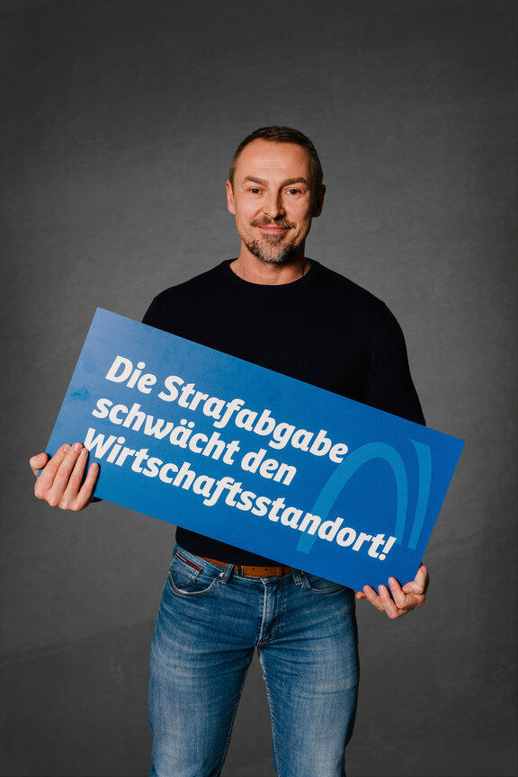 Eine Person steht vor einem neutralen grauen Hintergrund und hält ein großes blaues Schild mit der weißen Aufschrift: "Die Strafabgabe schwächt den Wirtschaftsstandort!"