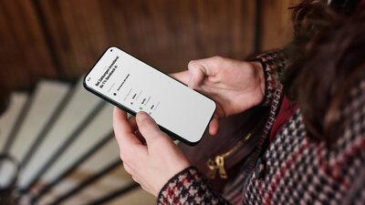 Zu sehen sind zwei Hände, die ein Smartphone halten. Auf dem Bildschirm ist die App von Trade Republic geöffnet, darin die Funktion Saveback und Sparpläne für Aktien von Adidas, Spotify und Meta.