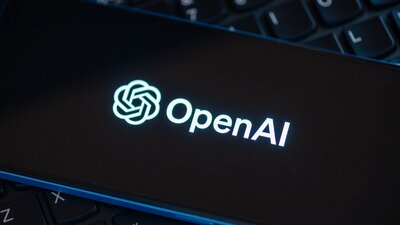Zu sehen ist ein auf einer Tastatur liegendes Handy, auf dessen Bildschirm das Logo der Firma OpenAI zu sehen ist.
