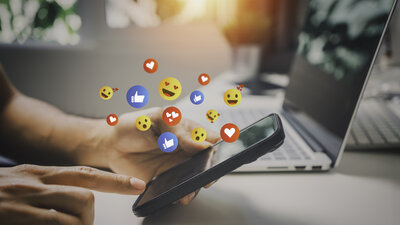 Hand hält Smartphone, Finger tippt auf Bildschirm, darüber schweben Symbole für soziale Medien wie Daumen hoch, Herz und lachende Emojis