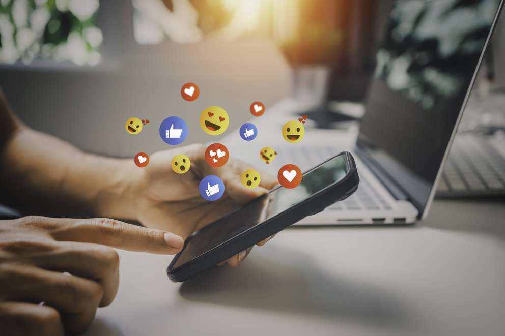 Hand hält Smartphone, Finger tippt auf Bildschirm, darüber schweben Symbole für soziale Medien wie Daumen hoch, Herz und lachende Emojis