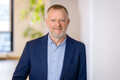 Mann mit kurzem Haar trägt ein blaues Sakko und ein hellblaues Hemd, steht an einer weißen Wand in einem hellen Raum.