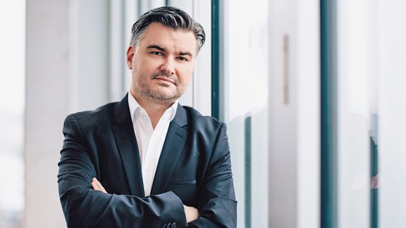 Mann mit dunklem Anzug und weißem Hemd, verschränkte Arme, steht vor großen Fenstern, selbstbewusster Gesichtsausdruck