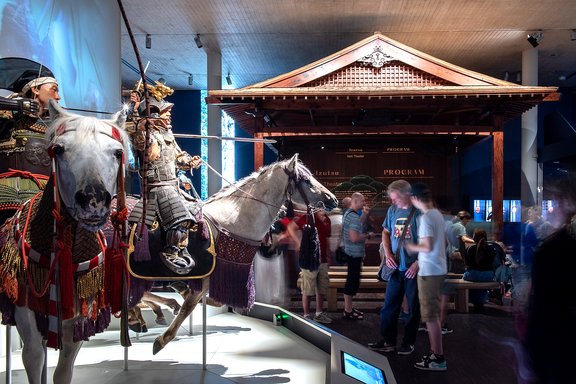 Samurai-Rüstung auf Pferd mit rotem Sattelzeug in Museumsausstellung, Besucher stehen und sitzen im Hintergrund vor traditionellem japanischem Gebäude