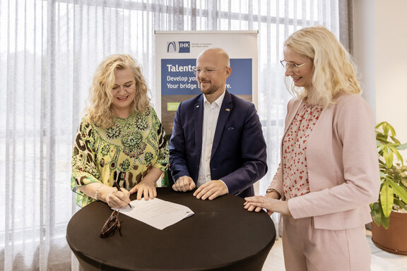 Drei Personen stehen an einem Stehtisch, auf dem ein Dokument liegt. Eine Person unterschreibt das Dokument, während zwei weitere danebenstehen. Im Hintergrund befindet sich ein Talentsbridge‑Banner.