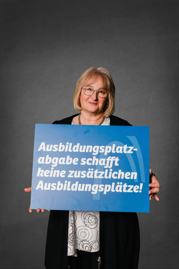 Eine Person steht vor einem neutralen grauen Hintergrund und hält ein großes blaues Schild mit der weißen Aufschrift: "Ausbildungsplatzabgabe schafft keine zusätzlichen Ausbildungsplätze!"