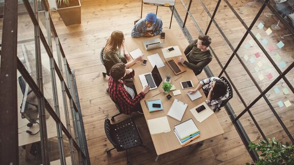 Zu sehen ist ein Büroraum, in der Mitte ei Tisch, an dem 4 Personen mit Laptops und Unterlagen sitzen und sich besprechen.