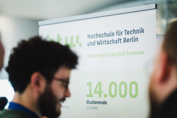 Zu sehen ist der verschwommene Kopf eines Mannes mit kurzen dunklen Haaren, Brille und Vollbart. Im Hintergrund steht ein Aufsteller der HTW Berlin - der Hochschule für Wirtschaft und Technik. Auf dem Austeller ist zu sehen, dass die Hochschule von 14.000 Studierenden besucht wird.