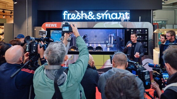 Mehrere Menschen mit Kamera fotografieren den KI-Roboter "Freh & Smart" der Circus Group in einem Rewe-Markt.