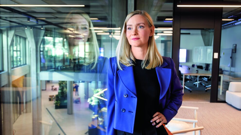 Das Bild zeigt eine blonde Frau im blauen Jackett in einem Büro, es ist Aiga Senftleben