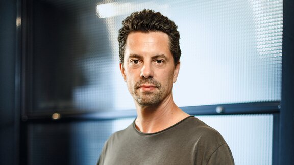 Zu sehen ist ein junger Mann, mit kurzen dunklen Haaren und einem grünbraunem Oberteil. Er schaut neutral in die Kamera.